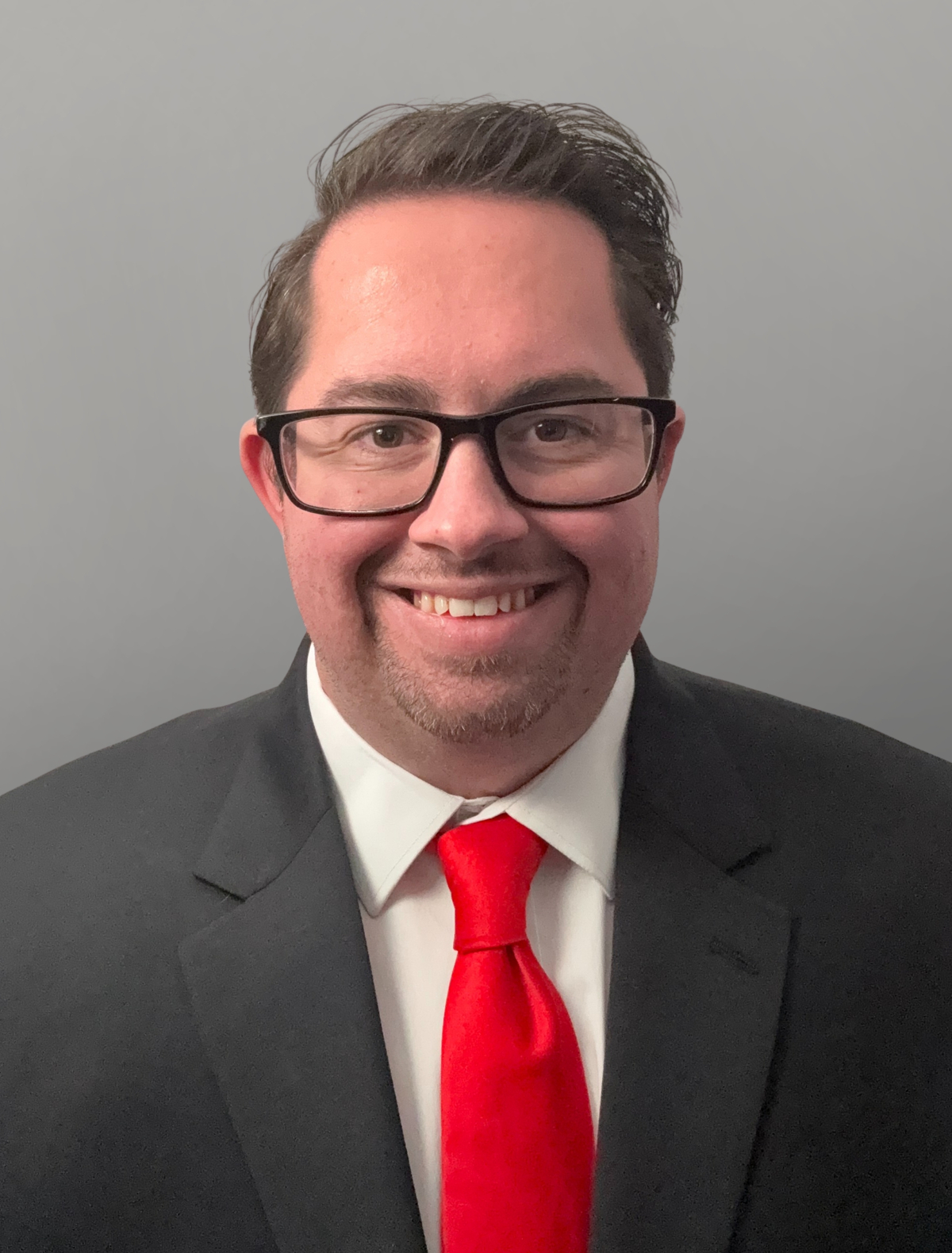 A man wearing glasses, a white shirt, a dark suit, and a red tie. 